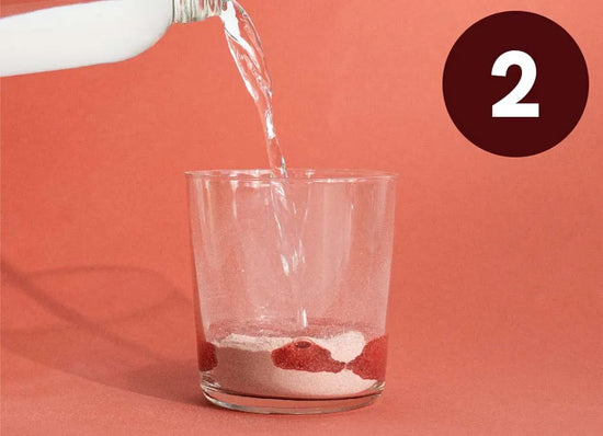 Vand hældes i et glas med pulver i bunden mod en rosa baggrund